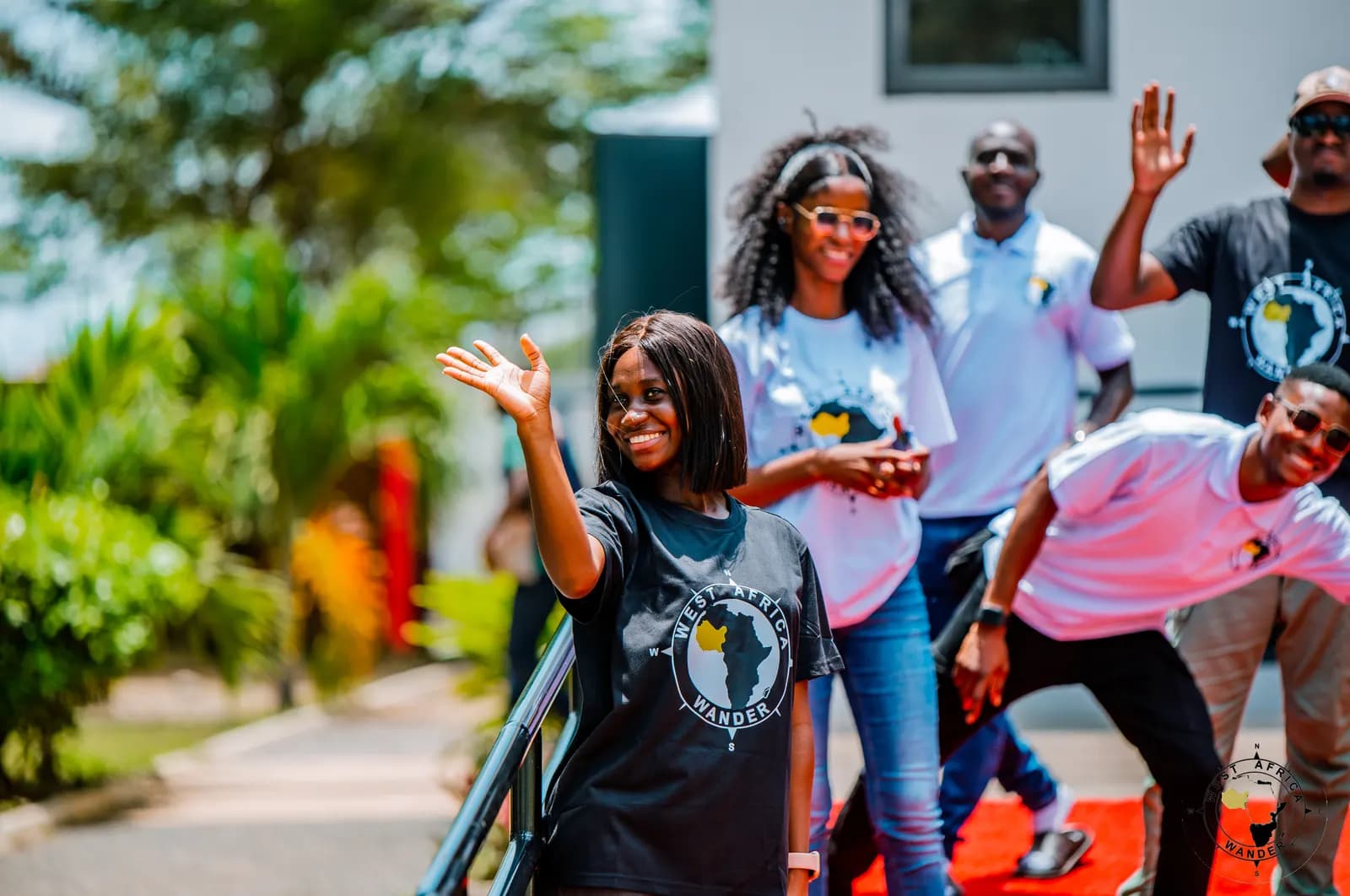 A West Africa Wander group in matching T-shirts posing and waving on a red carpet outside a white building