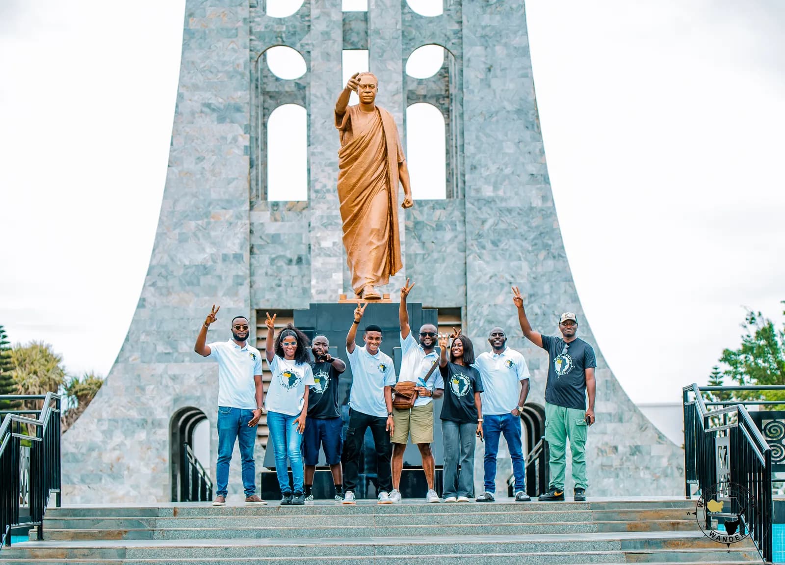 A West Africa Wander group raising their hands in front of a tall stone monument with a bronze statue