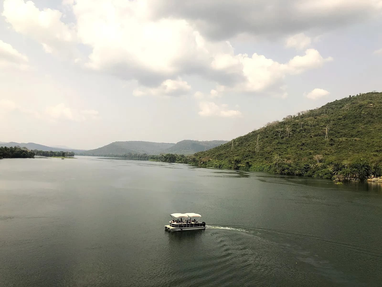 A covered passenger boat crossing calm water on the Volta between green hills