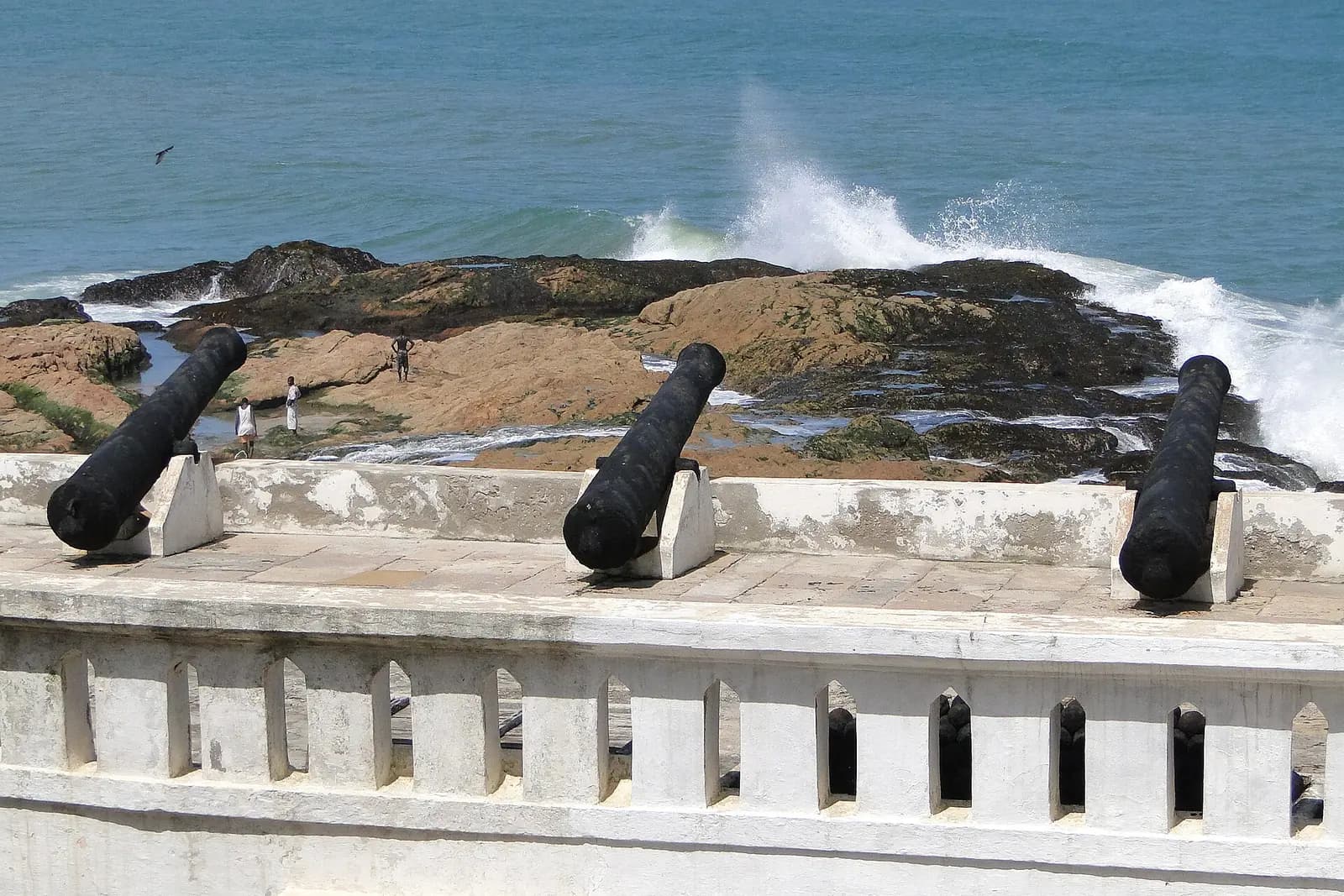 Three black cannons on the white sea wall of Cape Coast Castle, with waves breaking over the rocks below