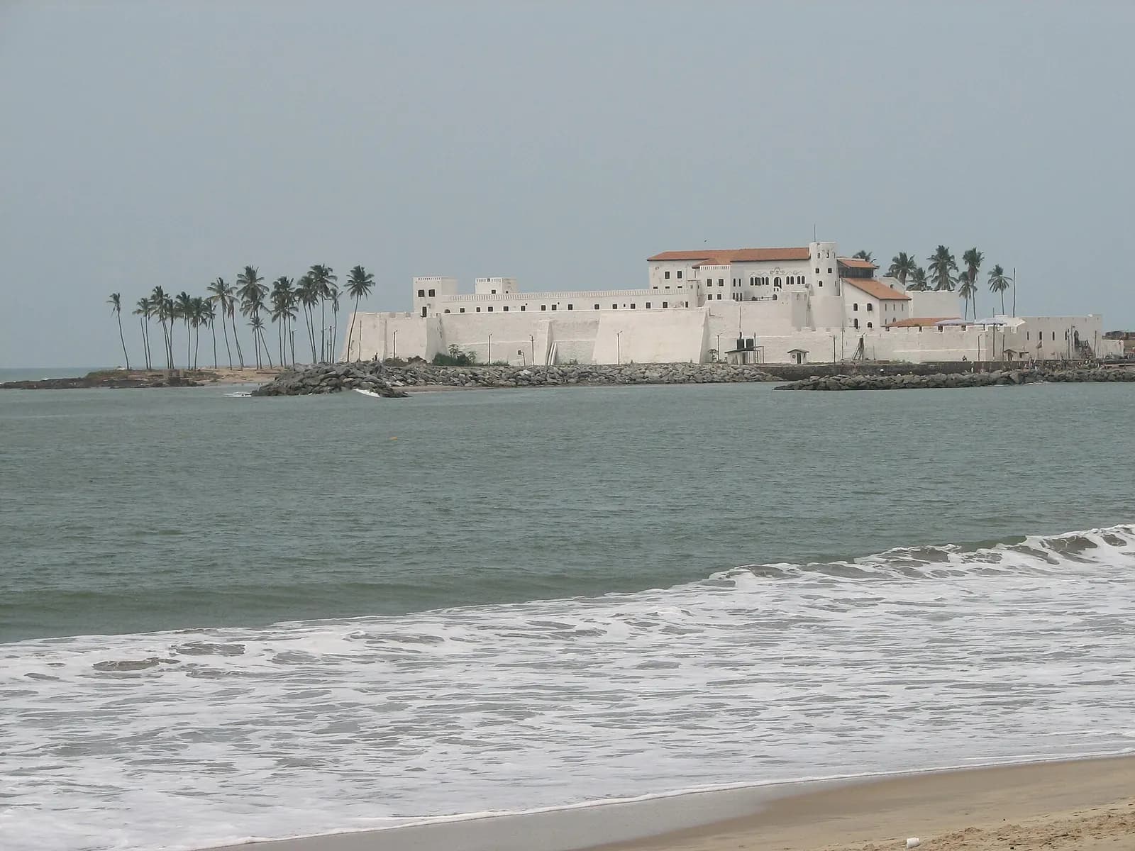 Elmina Castle's white walls and palm trees seen across the water from the beach
