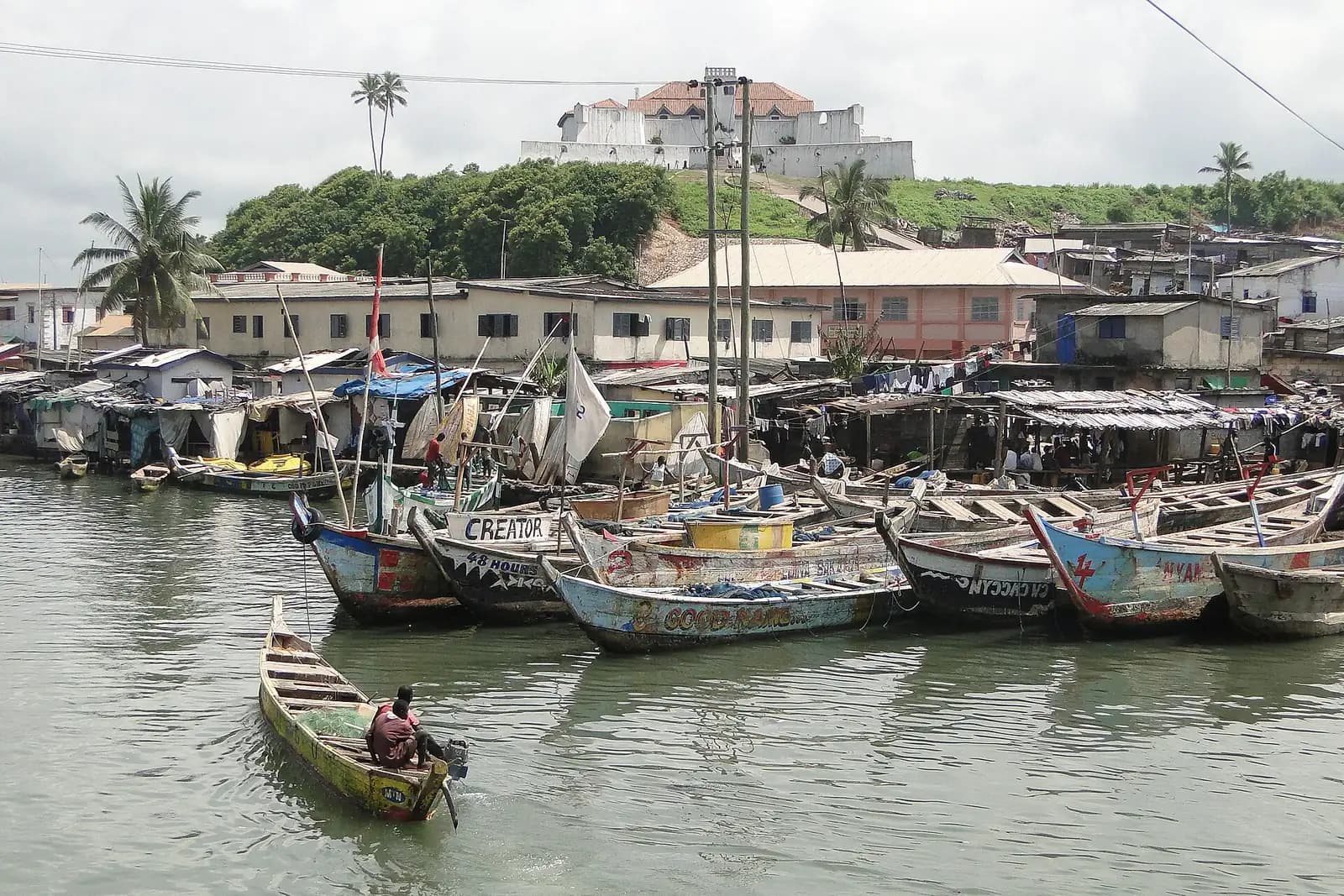 Painted wooden fishing canoes moored in Elmina harbour, with Fort St Jago on the hill above